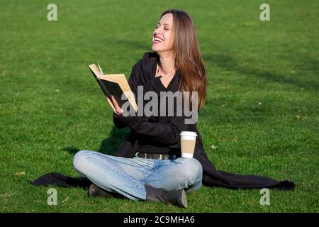 Brunette Mädchen sitzt gekreuzt auf einem grünen Rasen und lacht freudig beim Lesen eines schwarzen Hardback Buch mit einem Öko Papier Kaffeeglas. Stockfoto