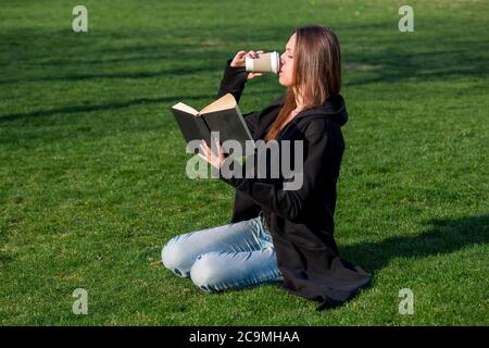 Ein junges Mädchen mit langen Haaren trinkt Kaffee aus einer Papiertasse und liest ein schwarzes Hardback-Buch, das an einem sonnigen Tag auf dem grünen Gras im Park sitzt Stockfoto