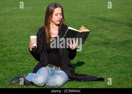 Ein Mädchen in einem schwarzen Mantel sitzt auf einem Rasen aus grünem Gras, hält eine Einweg-Papiertasse Kaffee in der Hand, und hält ein Hardback schwarzes Buch in ihrem anderen Stockfoto