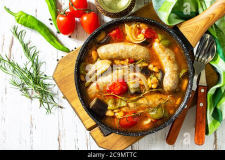 Bohnen, Gemüse und Räucherwürste mit Tomatensauce in der Pfanne auf dem Holztisch einkochen. Draufsicht flach lagen Hintergrund. Stockfoto