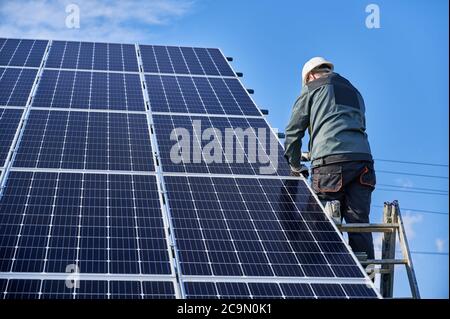 Rückansicht des Manntechnikers, der auf der Leiter steht und Photovoltaik-Solarpanel installiert. Männlicher Arbeiter mit Schutzhelm. Konzept der alternativen Energie und Energie nachhaltige Ressourcen. Stockfoto