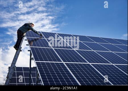 Männlicher Arbeiter in weißen Schutzhelm auf Leiter stehen und Reparatur Photovoltaik-Solarpanel-Station unter schönen blauen bewölkten Himmel. Konzept der alternativen Energie und Energie nachhaltige Ressourcen. Stockfoto