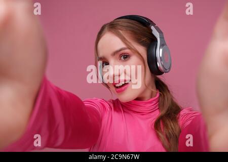 Nahaufnahme Porträt einer lächelnden jungen Rotschopf Frau machen Selfie mit zwei Händen. Lustige lächelnde Mädchen tragen Kopfhörer geben ein Geschenk isoliert auf rosa Stockfoto