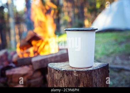 Nahaufnahme von weißem Campingbecher, auf Holzstumpf, vor Lagerfeuer und Schlafzelt. Stockfoto