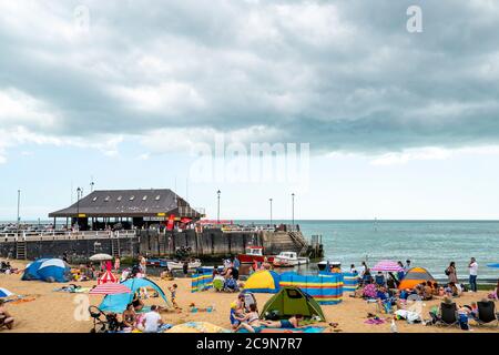 Leute, die am Strand sonnenbaden, aber versuchen, soziale Distanzierung im Sommer 2020 bei Broadstairs, an der englischen Kent-Küste, zu beobachten. Menschen am Strand mit dem Hafen im Hintergrund und grauen Wolken bilden sich darüber. Stockfoto