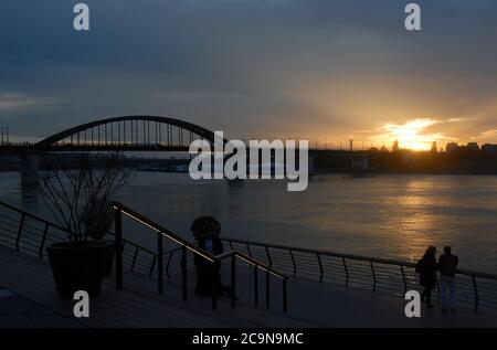 Die alte Sava-Brücke bei Sonnenuntergang in Belgrad, Serbien. Die Brücke überquert den Fluss Sava, wo ein Mann und eine Frau (nicht identifizierbar) den Sonnenuntergang beobachten Stockfoto