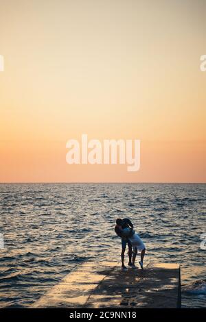 Silhouette eines Paares, das am Pier steht und küsst und den Sonnenaufgang am Strand im Sommer beobachtet Stockfoto