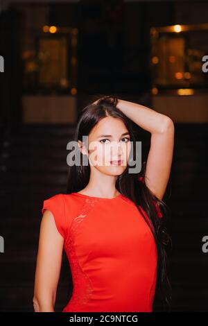 Porträt einer ziemlich schlanken Frau mit langen dunklen Haaren, trägt ein stilvolles langes rotes Kleid; sie hat eine Hand im Haar und schaut uns an Stockfoto