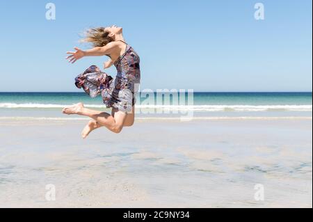 Blonde Frau mittleren Alters in einem grünen Kleid springen mit offenen Armen am Strand Stockfoto