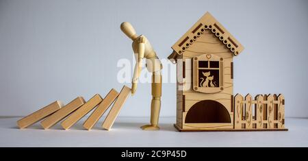 Holzmodell des Mannes, der hölzerne Domino-Blöcke in der Nähe des hölzernen kleinen Hauses hält. Stockfoto