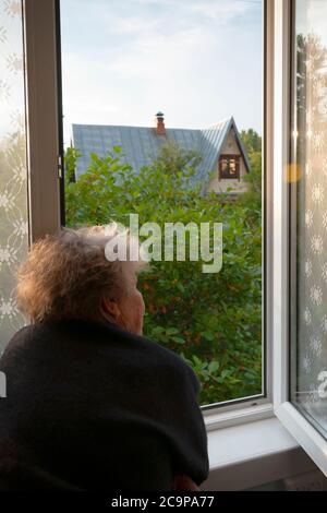 Ältere Frau mit Blick aus dem offenen Fenster des Landhauses Stockfoto