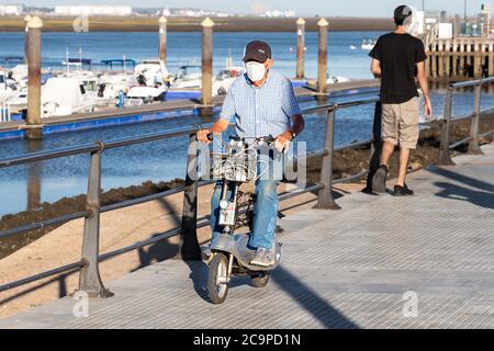 Punta Umbria, Huelva, Spanien - 10. Juli 2020: Ein älterer Mann, der mit einer Schutzmaske auf dem Bürgersteig einen Elektroroller fährt. Stockfoto