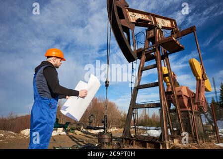 Seitenansicht des Ölarbeiterplans des Ölfeldes. Ingenieur in der Arbeit Uniform und Helm in der Nähe von Ölpumpe Jack unter schönen bewölkten Himmel stehen. Konzept der Erdölindustrie und Ölförderung. Stockfoto