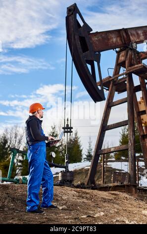 Vollständige Ansicht des Technikers mit Notebook und Blick auf die Kippmaschine der Ölpumpe. Männlicher Arbeiter in Uniform und Helm mit Laptop und Steuerung der Arbeit der Pumpe Jack. Konzept der Erdölindustrie. Stockfoto