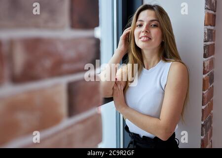 Porträt der jungen attraktiven kaukasischen Frau posiert vor der Kamera neben dem Fenster, sie schaut auf Kamera und lächeln. Schöne Frau mit langen Haaren steht ne Stockfoto