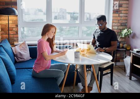 Alltag eines interracial Paar, junge Frau sitzen Blick auf Papier, während schwarzer Kerl sitzen arbeiten auf Laptop, freiberuflich während Quarantäne Stockfoto