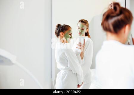 Lächelnde Freundinnen in Bademänteln und mit Lehmmasken im Gesicht im Badezimmer stehend und im Spiegel auf dem Smartphone fotografieren Stockfoto