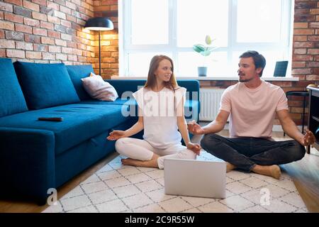Schwangere Frau praktiziert Yoga mit ihrem Mann zu Hause, sie benutzen Laptop, lernen neue Posen, sitzen in Lotushaltung, gehen zu meditieren Stockfoto
