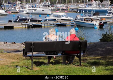 Nynashan, Schweden - 26. Juni 2020: Zwei ältere Frauen sitzen auf einer Parkbank mit der Marina im Hintergrund Stockfoto