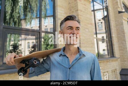 Заголовок Porträt von stilvollen reifen Mode-Modell tragen lässige Kleidung, posiert für Bilder. Glücklicher Skater mit Longboard, der auf der Straße läuft Stockfoto