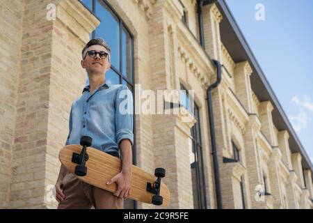 Glücklicher Skater mit Longboard, der auf der Straße läuft. Portrait von reifen Mode-Modell tragen lässige Kleidung und Sonnenbrille posiert für Bilder Stockfoto