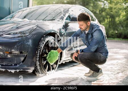 Luxus modernes Elektroauto in Seifenschaum im Freien bei Autowaschanlage. Seitenansicht des hübschen jungen kaukasischen Mann mit grünen Mikrofaser Auto Waschhandschuh Stockfoto