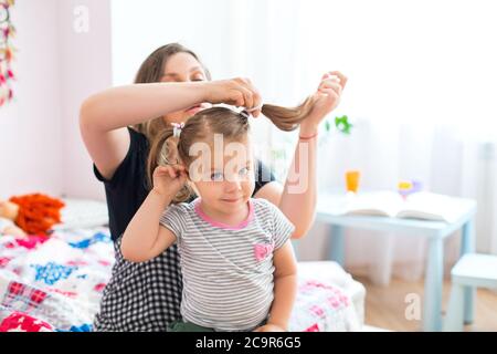 Crop Frau tun Pferdeschwänze für entzückende kleine Mädchen am Tisch sitzen und spielen mit Spielzeug während des Wochenendes zu Hause Stockfoto