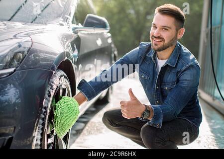 Lächelnder, hübscher junger Mann, der den Daumen nach oben zeigt und die Kamera anschaut, während er grünen Waschhandschuh verwendet, um die Felgen seines modernen Autos im Freien zu reinigen. Luxus Stockfoto