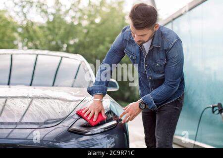 Junge gut aussehende kaukasischen Mann waschen und wischen seine modernen blau grauen Autoscheinwerfer an der Outdoor-Autowäsche Self-Service. Bärtiger Mann mit Rot Stockfoto