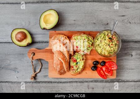 Flaches Lay Photo, frisch zubereitete Guacamole in kleiner Glasschüssel, Brot, Tomaten, Oliven am Arbeitstisch und zwei Avocados nebenan grauer Holzschreibtisch. Stockfoto