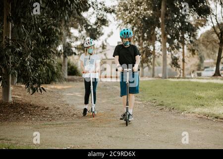 Zwei kaukasischen Kinder reiten Roller mit Masken während der Corona COVID-19 Pandemie Stockfoto
