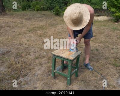 Der Mann in Stroh hatworking mit eletrischen Schleifmaschine, Werkzeug zum Schleifen und Polieren der Oberfläche, Renovieren und Polieren alten hölzernen kleinen Tisch Stockfoto