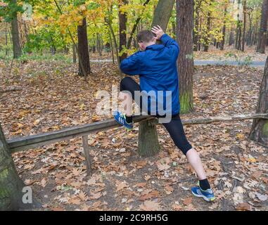 Junger Athlet Läufer Mann Stretching seine Beine in blauer Jacke und Schuhe in den Herbstwäldern, Aufwärmen bereit für das Joggen auf der Waldstraße draußen Stockfoto