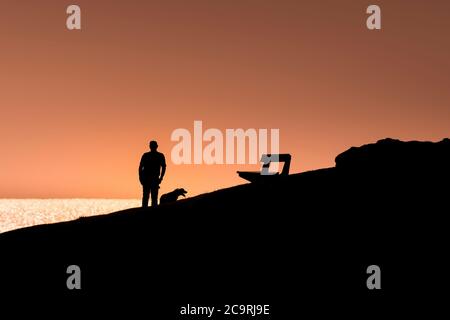 Ein Mann, der mit seinem Hund auf dem Pentire Point East läuft und auf das Meer blickt, das von einem intensiven, farbenfrohen Sonnenuntergang in Newquay in Cornwall umraunt wird. Stockfoto