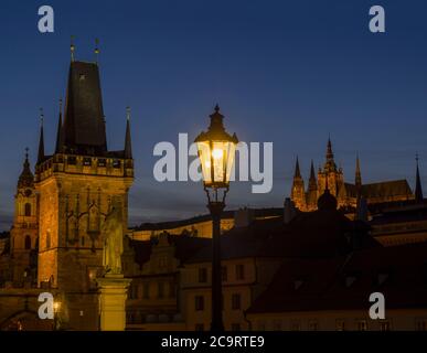 Blick von der Karlsbrücke auf den Mala Strana-Brückenturm mit glühender Straßenlampe nad Prager Burg bei Nacht, dunkelblauer Himmel. Stockfoto