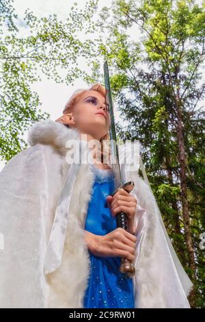 Die junge rothaarige Frau mit Elfenohren, blauem Kleid und weißem Umhang hält ein Schwert vor ihr Gesicht. Stockfoto
