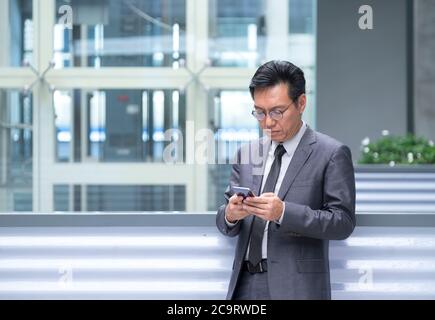Asiatischer chinesischer Geschäftsmann in Anzug und Krawatte, stehend und mit seinem Handy für SMS Stockfoto