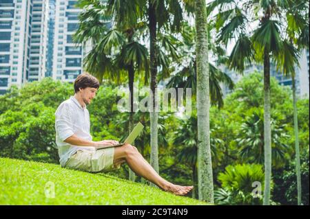 Mann Geschäftsmann oder Student in legerer Kleidung mit Laptop in einem tropischen Park auf dem Hintergrund von Wolkenkratzern. Dressing in einem weißen Hemd, beige Shorts Stockfoto