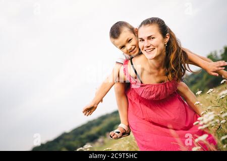 Frau spielt mit kleinen Sohn auf dem Feld mit Unkraut und Wiesenblumen am Sommertag. Sorglose Mutter, die Spaß mit Kind auf Schultern in Umarmung auf Stockfoto