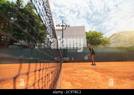 Weitwinkelansicht einer jungen schlanken Biracial-Frau mit einem Schläger in den Händen, der auf einem Sandplatz steht und eine Tennisrunde oder ein Warm-up hat Stockfoto