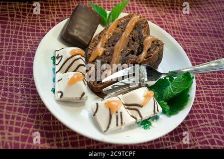 Portion Schokoladenkuchen mit Bonbons, die mit Minze, Toffee, Karamell und Puderzucker auf einer burgunderroten Tischdecke dekoriert sind Stockfoto