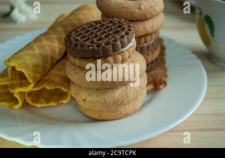 Waffelrollen und Kekse auf einem weißen Teller. Komposition auf einem hölzernen Hintergrund. Sichtbare Holzstruktur, Waffelstruktur Stockfoto