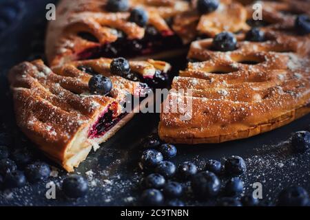 Hausgemachter süßer köstlicher Kuchen mit Heidelbeermarmelade, von dem ein Stück abgeschnitten wird und auf einem Backblech zwischen reifen Heidelbeeren liegt. Hausgemachter Kuchen. Stockfoto