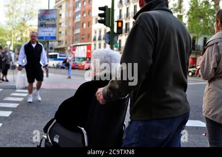 Madrid, Spanien - 20. April 2013: Ältere Dame im Rollstuhl, die mit Hilfe ihres Gefährten einen Fußgängerüberweg überquert. Stockfoto