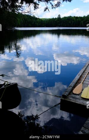 Sommer in der Hütte - kleiner See in Quebec, der den blauen Himmel mit flauschigen Wolken reflektiert - Kanu, Paddel und Dock im Vordergrund. Stockfoto