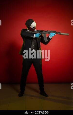Schauspieler Mann in einer Maske mit einer Waffe in den Händen posiert auf einem roten Hintergrund Stockfoto
