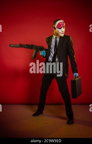Schauspieler Mann in einer Maske mit einer Waffe in den Händen posiert auf einem roten Hintergrund Stockfoto