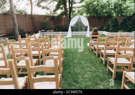 Hochzeitszeremonie im Stil von Rustikal. Holzstühle und ein weißer Bogen. Auf dem Stuhl sitzt der erste Gast und wartet auf den Anfang. Stockfoto