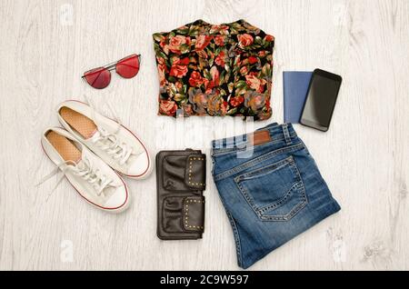 Shirt in Blumen, Brille, Turnschuhe, Jeans, Telefon und Reisepass. Holzhintergrund. Modisches Konzept, Draufsicht Stockfoto