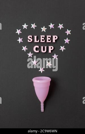 Rosa Menstruationstasse auf schwarzem Hintergrund mit Herz und Worten Schlaftasse. Konzept Möglichkeit der Nutzung Tasse in der Nacht, Null Abfall, Einsparungen, Minimalismus. Stockfoto